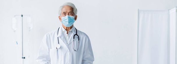 Iäkäs aasialainen lääkäri lääketieteellisessä naamiossa ja valkoisessa takissa katselemassa kameraa klinikalla, banneri  - Valokuva, kuva