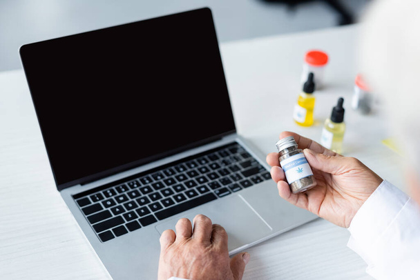 Ausgeschnittene Ansicht eines Arztes, der medizinisches Cannabis in der Nähe von Laptop und cbd-Öl hält  - Foto, Bild