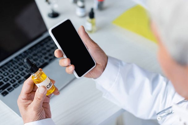 Blurred doctor holding smartphone and cbd oil near laptop in hospital  - Foto, Imagem