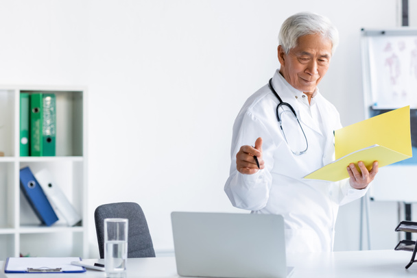 Senior doctor with pen and paper folder looking at computer near water and clipboard in clinic  - Foto, Bild