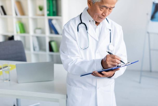 Elderly asian doctor writing on clipboard near blurred laptop in clinic  - Photo, image