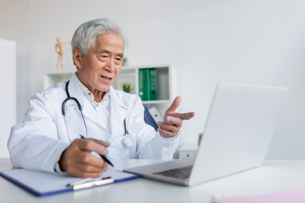 Elderly asian doctor pointing at laptop during video call and writing on clipboard in clinic  - Foto, afbeelding