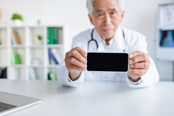 Smartphone met blanco scherm in handen van wazig aziatische arts in het ziekenhuis  - Foto, afbeelding