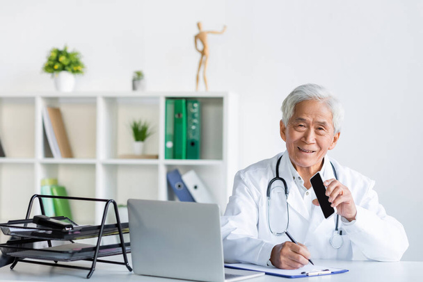 Uśmiechnięty azjatycki lekarz trzymając smartfona z pustym ekranem podczas pisania na schowku w pobliżu laptopa w biurze  - Zdjęcie, obraz
