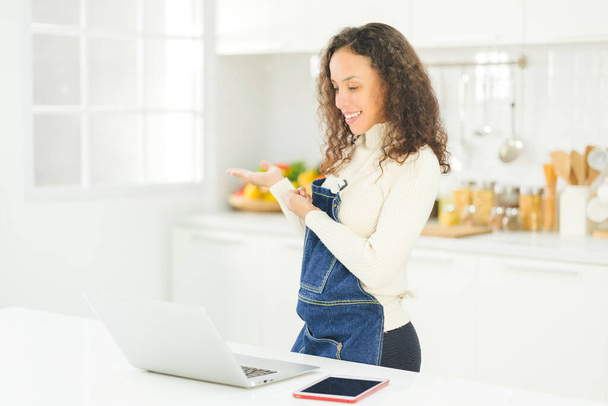 Portret van mooie jonge zwarte vrouw is live streaming koken met laptop in de heldere witte keuken. vrouw of huisvrouw bereidt zich voor om gezond voedsel voor het gezin te koken. Blijf thuis.. - Foto, afbeelding