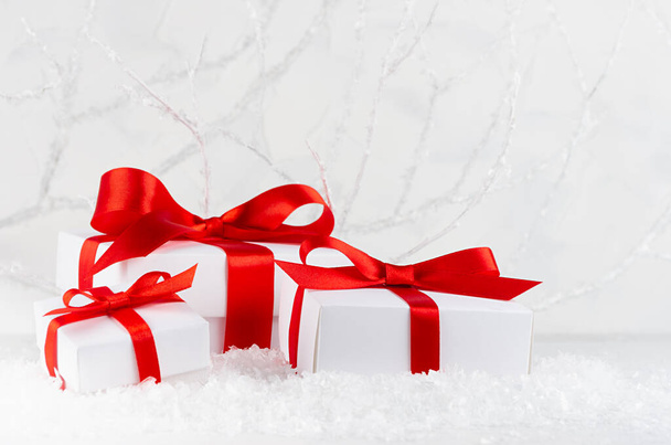Three bright white festive present boxes with red satin ribbon in decorative winter forest with frosty branches and snow, copy space. Christmas background with holiday packaging for celebration. - 写真・画像