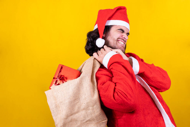Un bel giovanotto è vestito da Babbo Natale con un sacco pieno di regali. Babbo Natale è piegato indietro dal peso del pacchetto e ha un viso stanco. - Foto, immagini