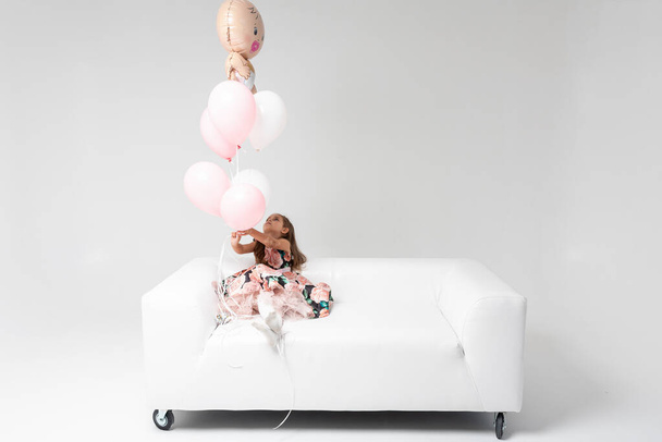 Little beautiful girl in a colored dress with blond hair sitting on a white sofa in the hands of colored balls holding, on a light background. She is smiling. Birthday, celebration - Fotoğraf, Görsel