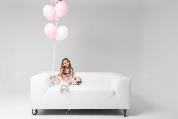 Little beautiful emotional girl in a colored dress with blond hair sitting on a white sofa in the hands of colored balls holding, on a light background. She is smiling. Birthday, celebration. - Photo, image