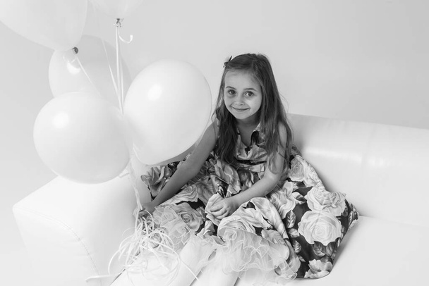Fekete-fehér fotó. kis gyönyörű érzelmi lány, színes ruhában, szőke hajjal, fehér kanapén ülve, színes golyók kezében, könnyű háttérrel. Születésnap, ünnep. - Fotó, kép