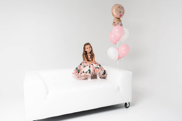 Little beautiful emotional girl in a colored dress with blond hair sitting on a white sofa in the hands of colored balls holding, on a light background. She is smiling. Birthday, celebration. - Фото, зображення