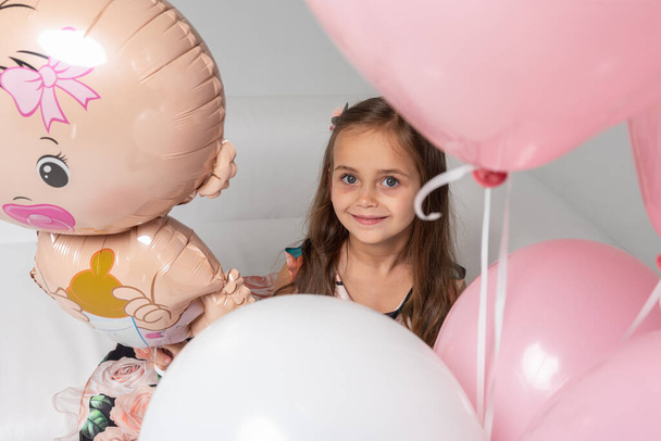 top view, little beautiful emotional girl in a colored dress with blond hair sitting on a white sofa between colored balls on a light background. She is smiling. Birthday, celebration. - Photo, Image