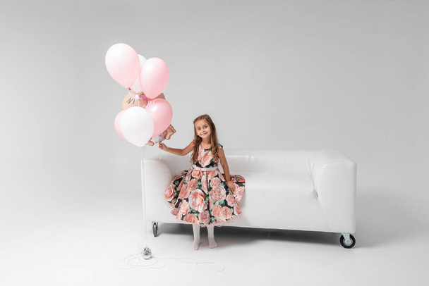 Little beautiful emotional girl in a colored dress with blond hair sitting on a white sofa in the hands of colored balls holding, on a light background. She is smiling. Birthday, celebration. - Photo, image