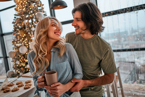 Couple casual stylish lovers are drinking coffee in the kitchen in new year morning. Young woman and her husband with a cups in them hands are smiling happy. Lifestyle christmas concept. - Foto, Bild