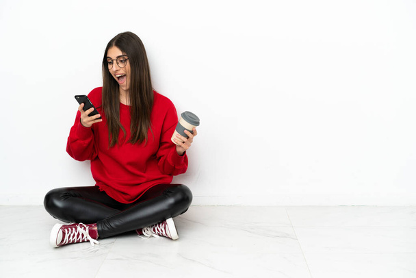 Mladá žena sedí na podlaze izolované na bílém pozadí drží kávu odnést a mobilní - Fotografie, Obrázek