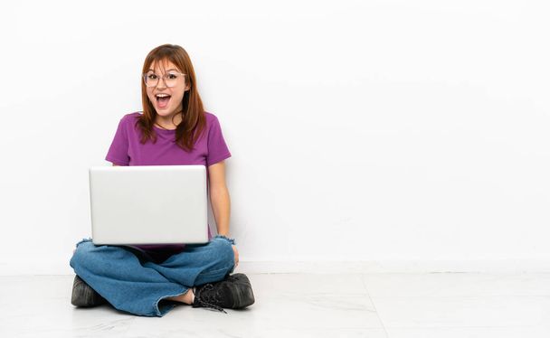 Dizüstü bilgisayarı olan kızıl saçlı kız yerde oturuyor ve yüzünde sürpriz bir ifade var. - Fotoğraf, Görsel