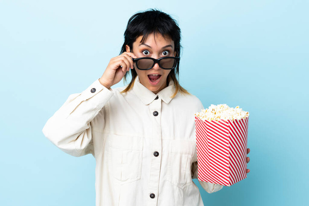 Mladá uruguayská žena na izolovaném modrém pozadí překvapena 3D brýlemi a drží velký vědro popcornů - Fotografie, Obrázek