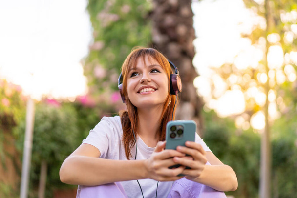 młoda ładna ruda kobieta na zewnątrz słuchanie muzyki z telefonu komórkowego i szczęśliwy - Zdjęcie, obraz