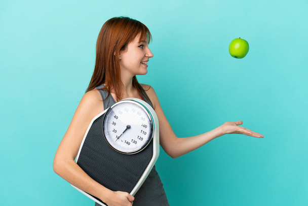 Redhead girl isolated on blue background with weighing machine and with an apple - Photo, Image