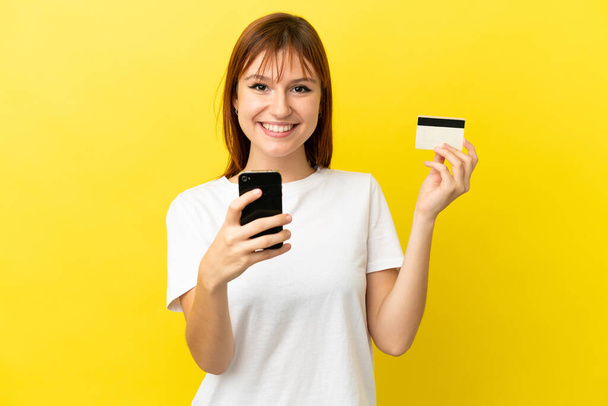 Рыжая девушка изолированы на желтом фоне покупки с мобильного телефона с помощью кредитной карты - Фото, изображение