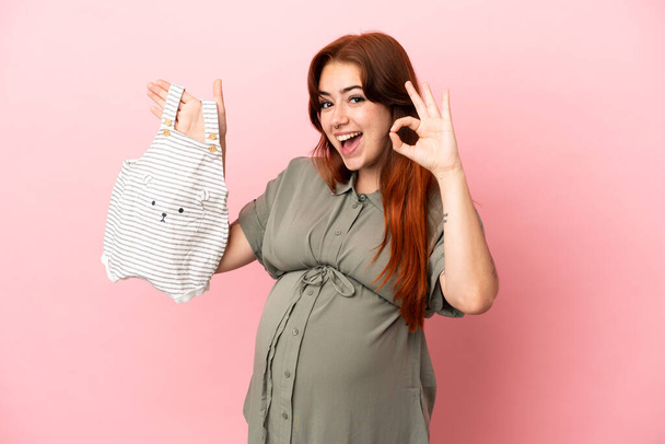 Genç kızıl saçlı beyaz kadın pembe arka planda izole edilmiş hamile ve üzerinde "Tamam" işareti olan bebek kıyafetleri taşıyor. - Fotoğraf, Görsel