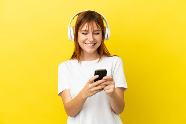 Ruda dziewczyna odizolowany na żółtym tle słuchanie muzyki z telefonu komórkowego i patrząc z przodu - Zdjęcie, obraz