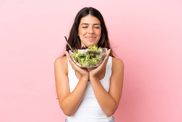 Pembe arka planda soyutlanmış beyaz bir kadın elinde mutlu bir ifadeyle bir kase salata tutuyor. - Fotoğraf, Görsel