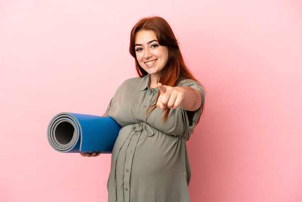 Νεαρή κοκκινομάλλα καυκάσια γυναίκα απομονωμένη σε ροζ φόντο έγκυος και πηγαίνει σε μαθήματα γιόγκα ενώ δείχνει μπροστά - Φωτογραφία, εικόνα