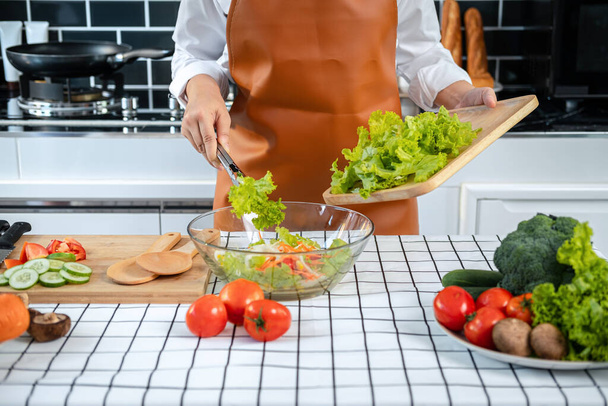 Asian housewife wearing apron and holding chopping board with lettuce to mixing with cucumber, tomato, carrot and spinach in bowl while cooking salad for healthy meal in her kitchen at home - Zdjęcie, obraz