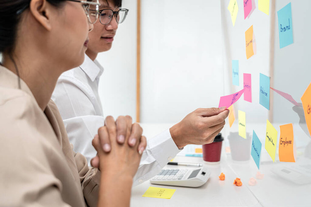 Businesswoman thoughtful about new startup project and brainstorming about planning marketing plan with partner while business man holding post it notes to share idea on a glass wall in office - 写真・画像