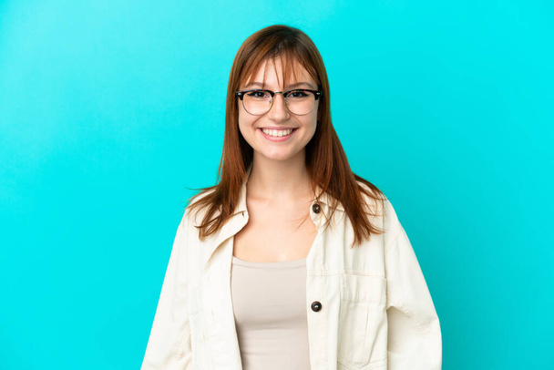 Redhead girl isolated on blue background With glasses with happy expression - 写真・画像