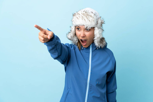 Young Uruguayan girl with winter hat isolated on blue background pointing away - Photo, image