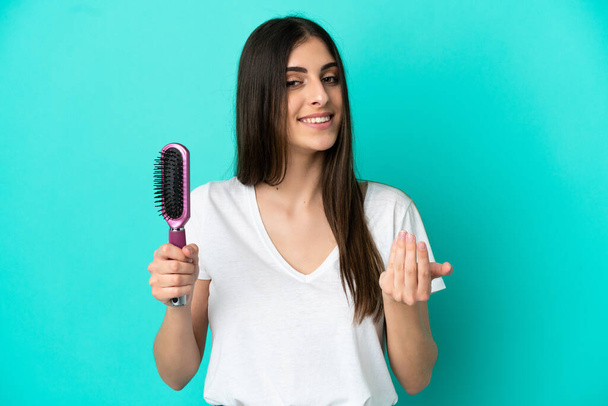 Young caucasian woman with hair comb isolated on blue background inviting to come with hand. Happy that you came - Foto, immagini