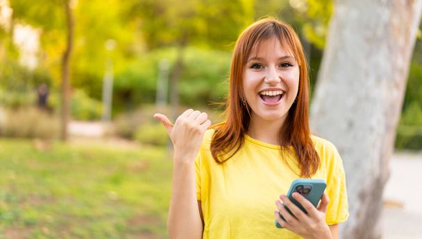 Young pretty redhead woman at outdoors using mobile phone and pointing to the lateral - Фото, изображение