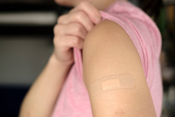 Χέρι µετά το εµβόλιο κατά του covid-19. Γυναίκα σηκώνει το μανίκι ενός t-shirt και δείχνει τον ώμο της με έναν επίδεσμο μετά τον εμβολιασμό. - Φωτογραφία, εικόνα