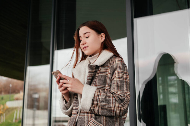 Молодая женщина в пальто стоит со смартфоном в руках на улице перед стеклянным окном. Работаю над мобильным устройством вне дома. Женщина-фрилансер, блогер. Тексты, чаты, чтение. - Фото, изображение