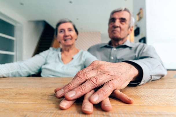 пожилая пара дома, сидящая за столом, держась за руку - сосредоточьтесь на руках на переднем плане - Фото, изображение