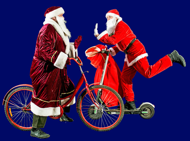 Két boldog Mikulás karácsonyi sapkában és piros öltönyben egy robogón és egy biciklin, tele ajándékokkal. Boldog új évet 2022-ben. Boldog karácsonyt! Fogalom. - Fotó, kép