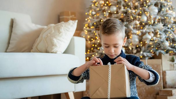 Το παιδί ανοίγει το Χριστουγεννιάτικο δώρο. Αγόρι κοντά στο χριστουγεννιάτικο δέντρο με κουτιά δώρων. Διακοσμημένο σαλόνι. Χριστουγεννιάτικη διάθεση. Παραμονή Πρωτοχρονιάς - Φωτογραφία, εικόνα