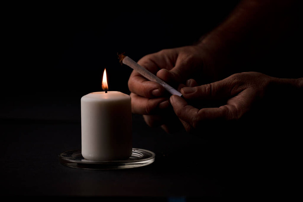 свеча зажжена пламенем, человеческие руки держат сигару, черный фон. (сфокусироваться на свечах). - Фото, изображение