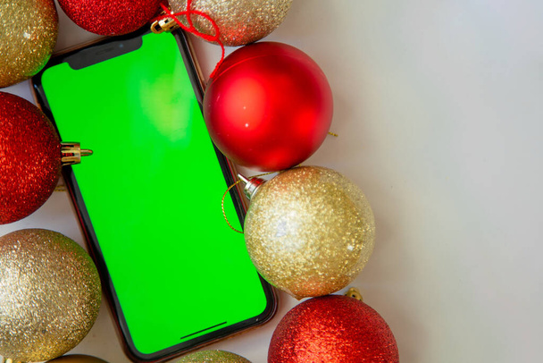 A phone with a green screen lies among the golden and red Christmas balls. - Foto, imagen