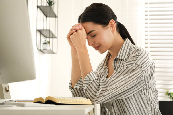 Beautiful young woman praying over Bible at desk - 写真・画像