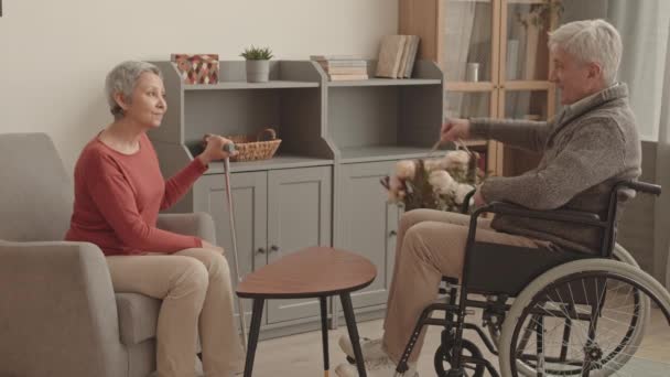 Volledig zijaanzicht van oudere blanke man in rolstoel geeft bloemen aan senior Aziatische vrouw met riet zitten in fauteuil in de woonkamer overdag - Video