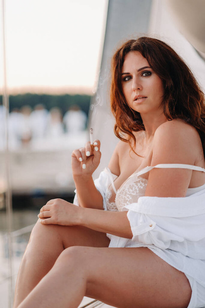 sexy woman in a white shirt with a cigarette in her hands enjoys the sunset on her private white yacht. - Zdjęcie, obraz