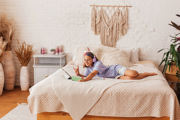 Beautiful teenager girl communicates online by phone while lying on the bed at home. Child in headphones learn online while lying on the bed - Foto, Bild