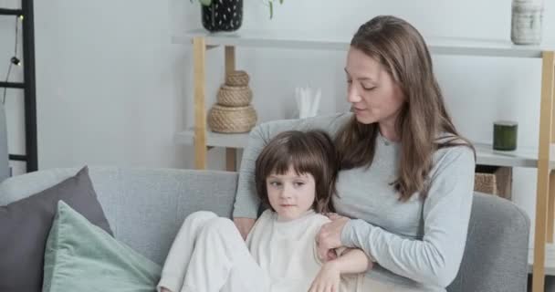 Fürsorgliche Mutter beruhigt sich und umarmt kleinen entzückenden Sohn, der zu Hause zusammen auf der Couch sitzt - Filmmaterial, Video