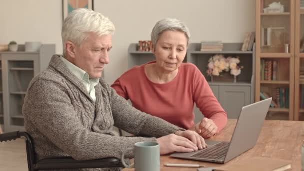 Waist-up of short-haired Asian woman and her senior Caucasian husband in wheelchair sitting at desk at home, man typing on portable computer, spouses talking - Footage, Video