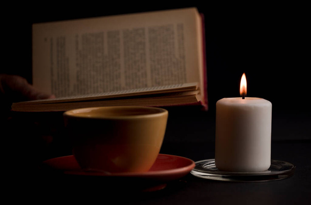 A candle lit with a flame, with a cup of tea with smoke, and a reading book in the background. (focus on candle). - Фото, изображение