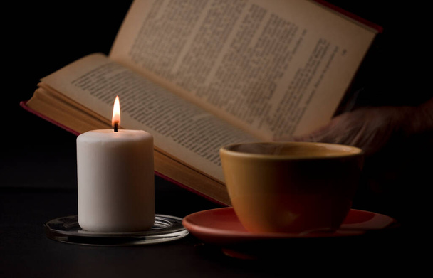 A candle lit with a flame, with a cup of tea with smoke, and a reading book in the background. (focus on candle). - Photo, image