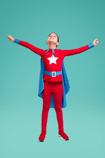 Happy kid in superhero costume standing in studio with outstretched arms - Фото, изображение